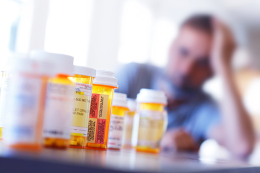 A large group of prescription medication bottles sit on a table in front of a distraught man who is leaning on his hand as he sits at his dining room table.  The image is photographed with a very shallow depth of field with the focus being on the pill bottles in the foreground.